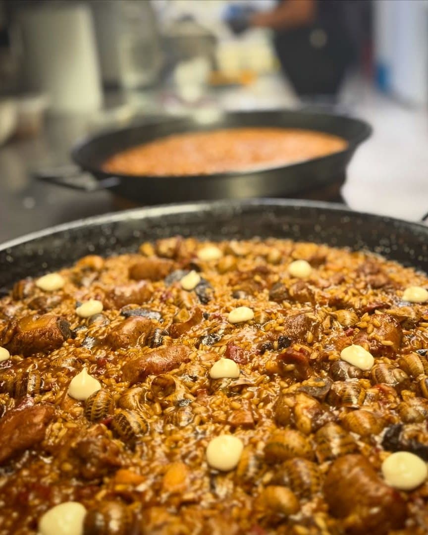 Primer pla d'una paella d'arròs amb caragols, trossos de carn i punts de salsa blanca, amb una altra paella desenfocada al fons.