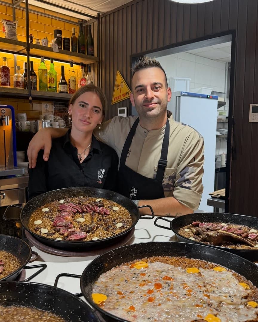 El xef Ramon Tudela abraçant una companya de l'equip, tots dos darrere de diverses paelles d'arròs acabades.