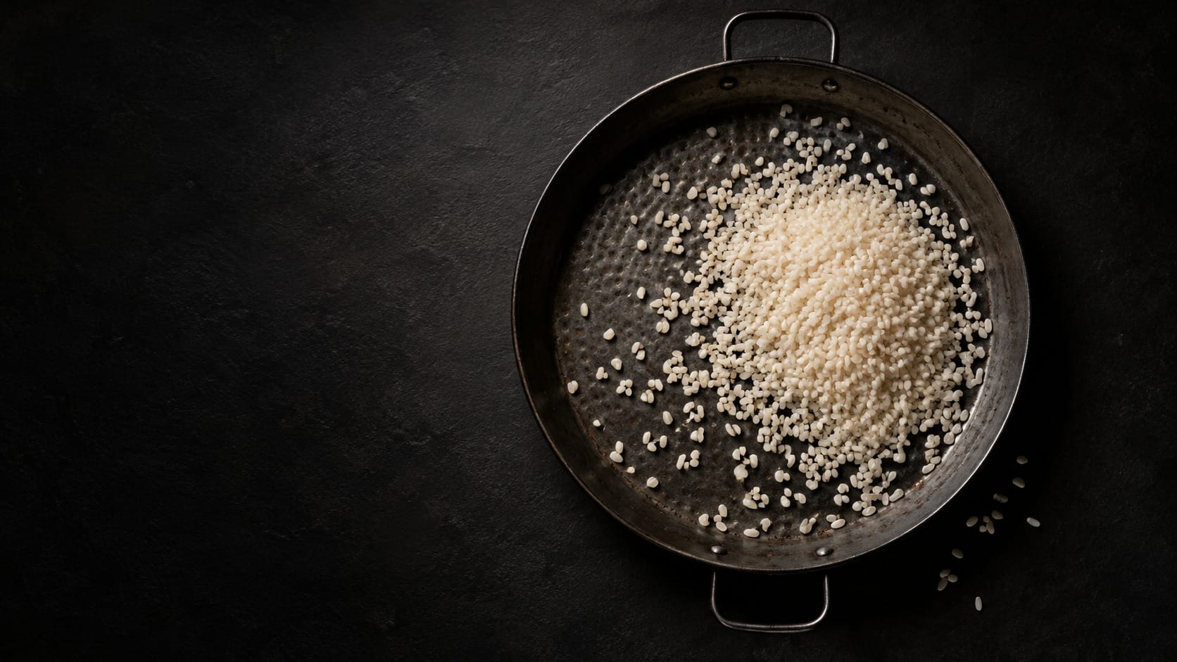 Imatge d'ambient creada amb IA: grans d'arròs cru amuntegats dins d'una paella negra, sense cap altre ingredient, vista des de dalt sobre un fons fosc.